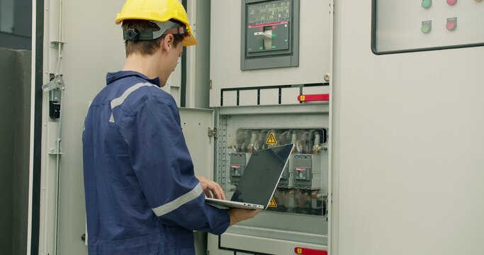 An electrical engineer performs a system diagnostic check with a laptop, conducting maintenance on an industrial power distribution control panel in a factory.