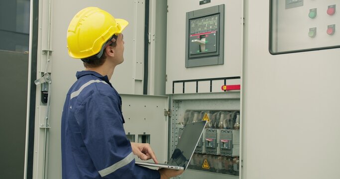 An electrical engineer performs a system diagnostic check with a laptop, conducting maintenance on an industrial power distribution control panel in a factory.