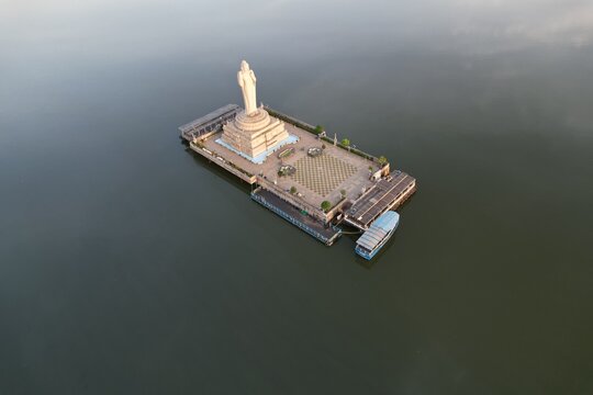 Aerial view of the monolithic Buddha statue standing on a platform in the middle of Hussain Sagar lake with a boat docked nearby in Hyderabad, Telangana, India.