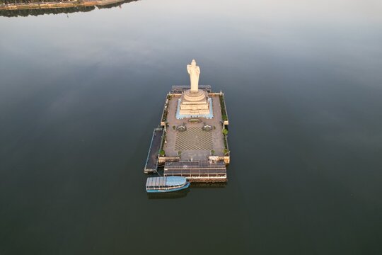 Aerial view of the monolithic Buddha statue on an island in Hussain Sagar lake, with a boat docked at the pier and calm water in Hyderabad, Telangana, India.