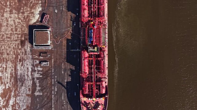 A red cargo ship is positioned at the industrial port next to the dock. Workers are engaged in loading and unloading goods from the vessel in broad daylight.
