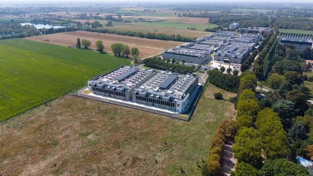 Aerial view of DATA4 MILAN CAMPUS, a large data center facility with industrial buildings and cooling units on roofs, surrounded by fields and trees in Zona Industriale, Lombardia, Italy.