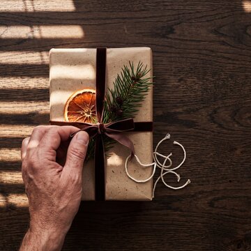 Hand of a person wrapping a rustic Christmas gift box with craft paper, velvet ribbon, dried orange, and fir branch on a wooden table