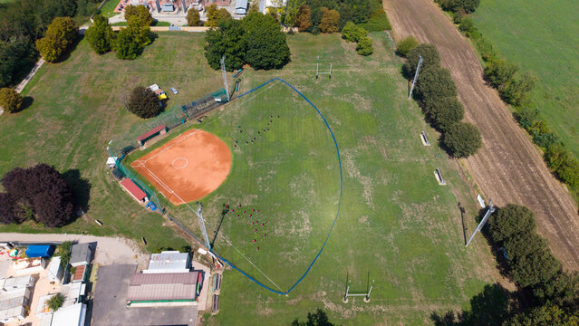 Aerial view of Campo da Baseball with players on the field, red clay infield, and green grass outfield surrounded by trees and rural landscape in Cornaredo, Lombardia, Italy.