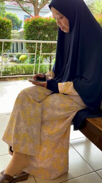 A young woman wearing a traditional hijab and floral pattern trousers sits calmly on a wooden bench in an outdoor garden setting, surrounded by greenery.