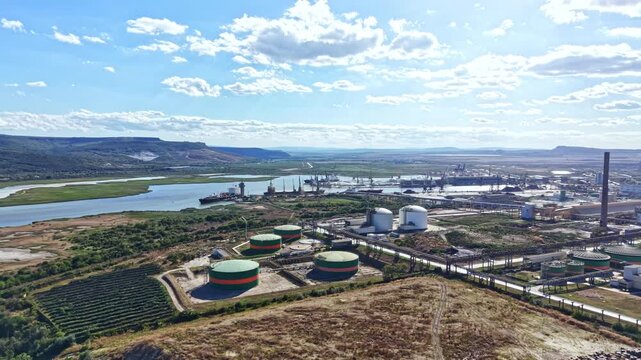 View of an oil refinery and storage tanks beside a river. Industrial activities are ongoing in the area. Green landscapes surround the site under a clear sky.
