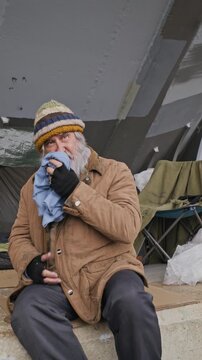 Portrait of senior Caucasian homeless man sitting near shelter, wiping face and looking at camera as lens zooms in. Clip fits poverty awareness and charity campaigns, vertical