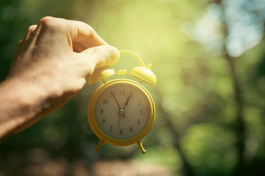 Hand holding yellow alarm clock with green bokeh nature background.