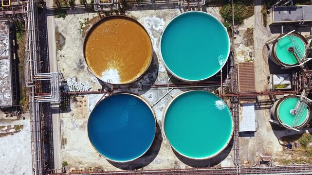 Large circular tanks filled with various liquids in bright colors are located at an industrial site. The scene shows different shades like blue, green, and brown under sunlight.