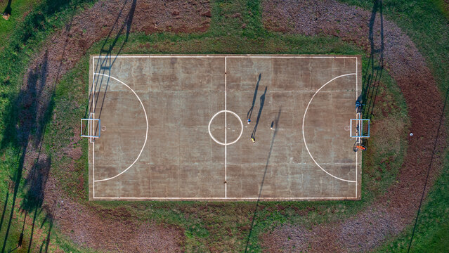 Aerial view of a concrete multi-sport court with people casting long shadows on the surface, surrounded by green grass in Posadas, Misiones Province, Argentina.