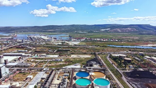 A view of an industrial area featuring large water storage tanks, port facilities, and various buildings under a clear blue sky. Mountains are visible in the background.
