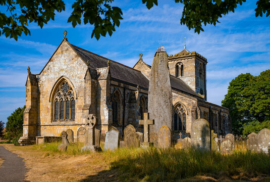 Historic All Saints&rsquo; Church in Rudston, East Yorkshire, with the remarkable prehistoric Rudston Monolith rising among old gravestones in the churchyard on a sunny summer day.