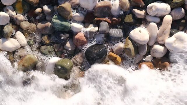 Close-up view of foamy water cascading over various pebbles on a beach, highlighting the textures and colors of the stones in the water