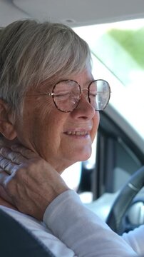 Elderly woman with glasses driving a car and experiencing sudden neck pain, massaging her tired and stiff neck muscles with her hand while sitting at the steering wheel in discomfort