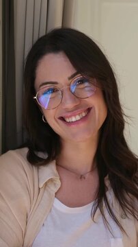 Close-up of a happy and beautiful brunette woman wearing glasses smiling at the camera, her face radiating joy and confidence with a genuine and warm expression in a cozy indoor setting