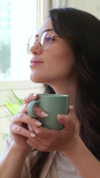 Calm young brunette woman wearing glasses holding a mug, enjoying a warm beverage like coffee or tea in the morning, peacefully looking out of a bright window and smiling in a cozy home