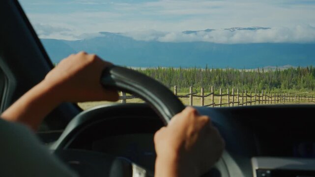 Driver grips steering wheel, eyes forward. Mountainous landscape stretches ahead. Road winds past wooden fence, vineyard rows. Clear blue sky, calm drive. Perfect for travel, adventure, road trip