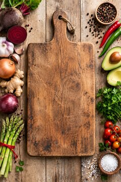 Wooden cutting board surrounded by fresh vegetables and herbs on rustic wooden table