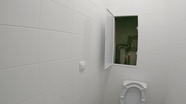 First person view of a small restroom with white wall tiles and light floor tiles. A pedestal sink and toilet are present. An open wall hatch reveals exposed plumbing pipes and valves