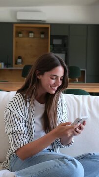 Young beautiful woman sitting on the couch checking her mobile phone and celebrating a victory or some good news she has just received, expressing happiness, joy, and excitement