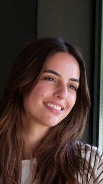 Cheerful young brunette woman with natural beauty and a radiant smile looking outside a window, enjoying the daylight and feeling optimistic about her future with a positive mindset