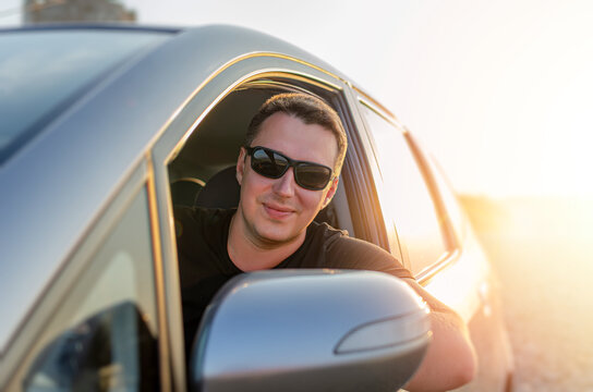 A smiling man wearing sunglasses looks out from the open driver window of his modern vehicle at sunset.