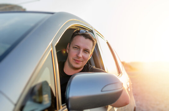 A smiling young man looks out from the open window of a modern car during a sunny road trip.