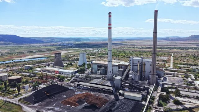 A coal power plant stands beside a river. Large smoke stacks release emissions into the air. The surrounding landscape shows open fields and mountains in the distance.