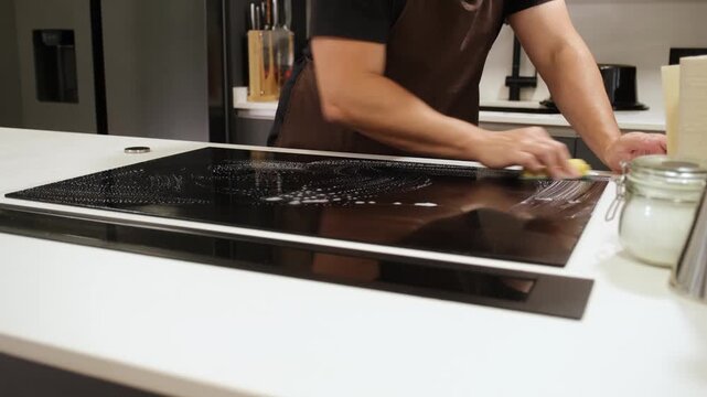 Diligent man in an apron wiping down a soapy glass ceramic stove in a modern kitchen