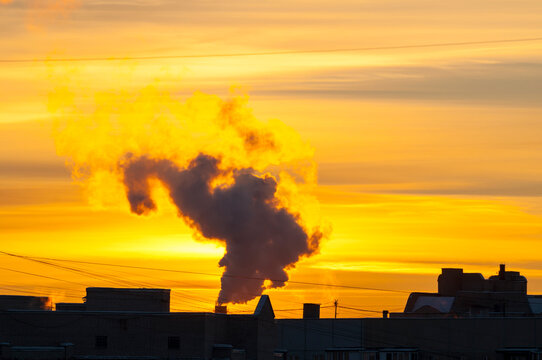 Industrial sunset background. A thick plume of smoke rising from a factory chimney. Bright golden sky and silhouetted rooftops at the horizon