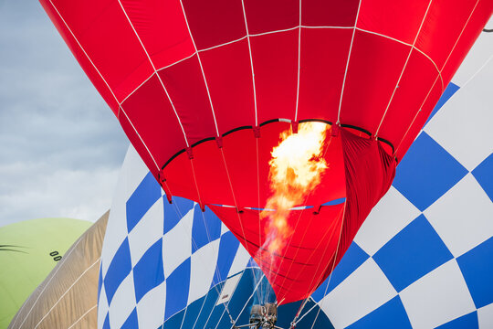 Powerful bright propane burner flame firing intense fire into opening of red hot air balloon envelope during inflation preparation at outdoor aeronautical festival event under cloudy blue sky backdrop