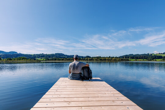 Back view of man with backpack sitting on wooden jetty over lake enjoying tranquil mountain view near Bernbeuren and Auerberg, Bavarian nature travel concept