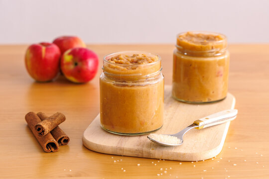 Homemade applesauce in glass jars on wooden board with fresh red apples cinnamon sticks and sugar spoon, autumn preserve and dessert ingredient concept