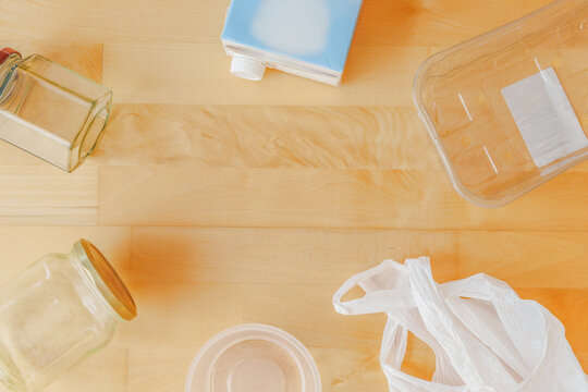 Top view flat lay of various household recyclable packaging materials like glass jars plastic containers beverage carton and plastic bag on light wooden background with central copy space