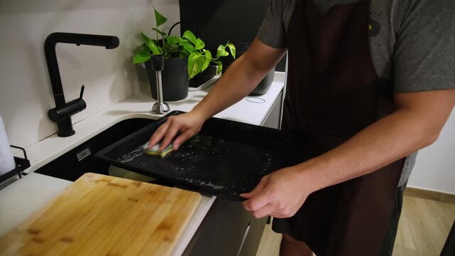 Diligent man wearing an apron thoroughly washing a dirty baking tray with a sponge in the kitchen