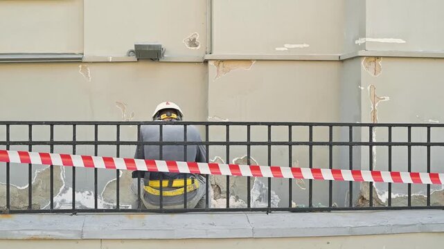 A construction site is shown with safety barriers marking off a work area. A worker wearing a helmet is present, along with building materials and unfinished walls visible in the background