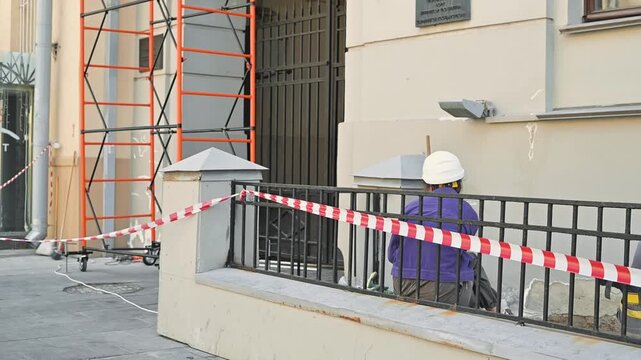 A construction worker in a helmet is seated near a fence with red-and-white safety tape. Scaffolding is visible in the background, indicating ongoing maintenance on the building