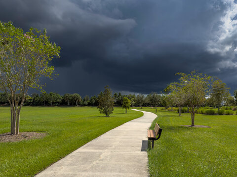 Dramatic view of a community park with sunny concrete sidewalk between green landscaped fields in the path of a thunderstorm on a summer afternoon in Florida. For motifs of imminent change.