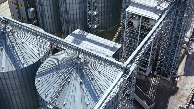 Grain silos are seen from above at a storage facility in an agricultural area. Transport equipment connects the silos for moving grain. Sunlight shines on the structure.