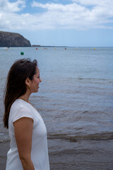 Woman in white stands by calm ocean, gazing thoughtfully toward horizon. Her relaxed posture suggests peaceful contemplation or quiet solitude © AlbertoRodriguez