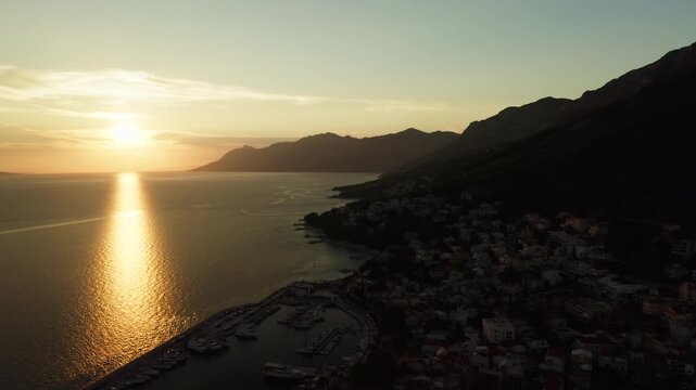 Bright golden sunset reflects on Adriatic sea water in Baska Voda Croatia. Warm sunlight illuminates mountains and coastal harbor. Tourists enjoy peaceful evening tranquility on vacation.