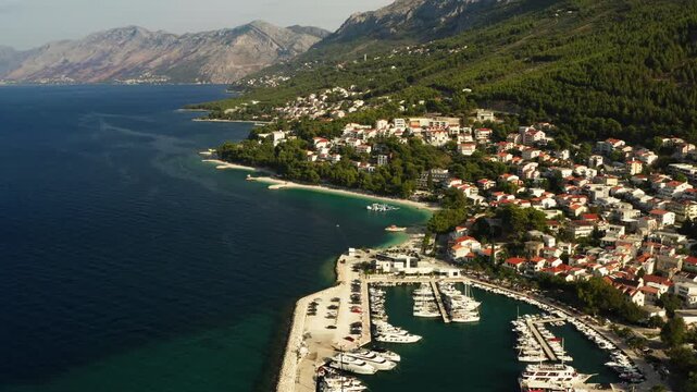 The Adriatic coastline stretches along the resort town of Baska Voda in Croatia. Biokovo mountains and pine hills border the blue sea. Beaches and a marina harbor line the resort waterfront.