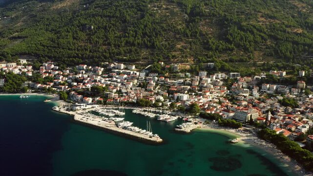 Baska Voda coastal resort town sits along Adriatic sea in Croatia. Mountain Biokovo rises above residential houses and harbor marina. Moored boats and yachts line stone piers in clear water.