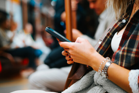 A person is focused on their smartphone while sitting on a subway train. Other commuters are visible in the background, some engaged in their own activities