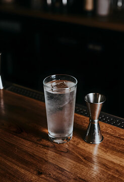 Hospitality and beverage service, chilled water glass with ice cube and metal jigger on wooden bar counter in dark restaurant or cocktail lounge