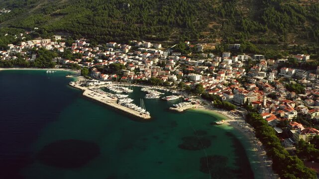 Baska Voda resort town borders the azure Adriatic Sea with a busy yacht marina. Terracotta roofs cover hillside houses along sunny pebble beaches. Scenic vacation getaway in Dalmatia, Croatia.