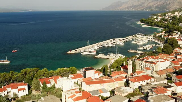 A passenger boat departs Baska Voda marina into the Adriatic Sea. Clear turquoise water surrounds the stone breakwater pier and beach. Coastal resort harbor bustles in Dalmatia, Croatia.