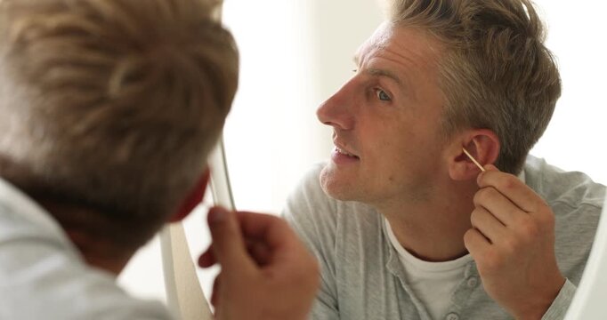 Man cleaning ear with cotton swab near mirror indoors