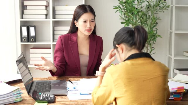 Angry asian female boss scolding a stressed employee in the office, pointing at documents and laptop with a frustrated expression for a work mistake and poor performance