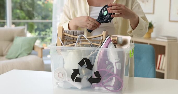 Recycling. Woman putting different broken household appliances into plastic container at white table indoors, closeup